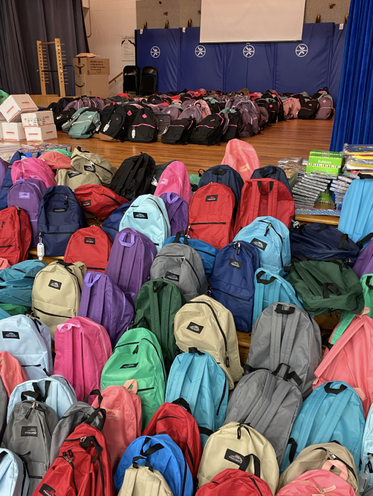 Over 50 individual backpacks of varying colors are arranged in front of an indoor wooden stage. On the stage are more backpacks clustered together. Blue mats line the stage and various boxes and school supplies are scattered around the stage.
