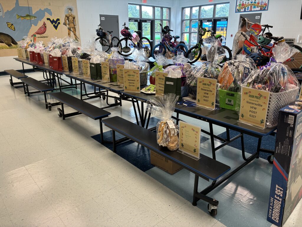 A school cafeteria features tables lined with wrapped gift baskets and prize boxes. Bikes are displayed in the background, creating a festive and charitable atmosphere.