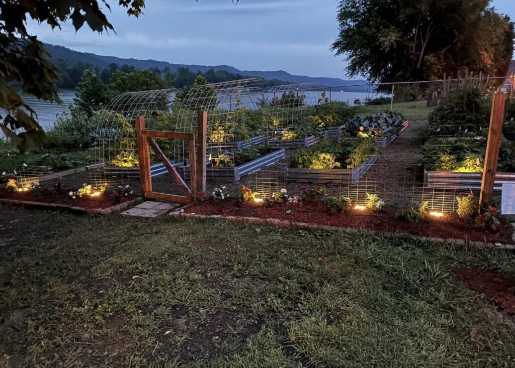 Illuminated vegetable garden with raised beds and wire arches at dusk, bordered by flowers. A serene river and hills are visible in the background.
